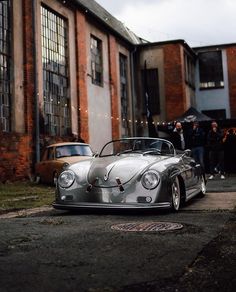 an old silver car parked in front of a brick building with people standing around it