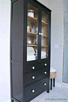 a black cabinet with glass doors and drawers in the corner is next to a white toilet