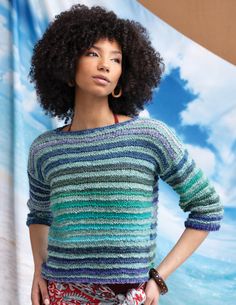 a woman with an afro standing in front of a blue sky and clouds backdrop wearing a multicolored sweater