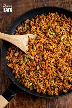 a skillet filled with rice and vegetables on top of a wooden table next to a wooden spoon