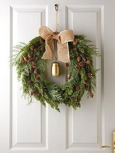 a christmas wreath hanging on the front door with a gold bell and pine cones tied to it