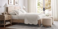 a bed with white sheets and pillows in a bedroom next to a table, chair, ottoman and window