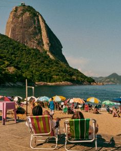 two people sitting in lawn chairs on the beach with umbrellas and mountains in the background