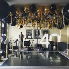 a room filled with balloons and black and gold streamers on the ceiling, along with a man standing in front of a stage set up for a party