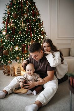 a man and woman are sitting in front of a christmas tree with a baby on their lap