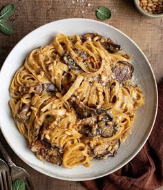 a white bowl filled with pasta and mushrooms on top of a wooden table next to silverware