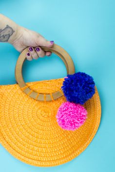 a woman's hand holding a yellow straw purse with pom - poms