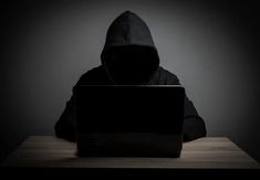 a hooded person sitting at a table with a laptop in front of him, looking down