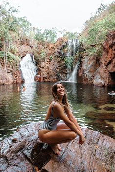 a beautiful woman sitting on top of a rock next to a body of water with a waterfall in the background