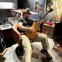 a man sitting in a chair with a guitar on his lap and wearing a hat