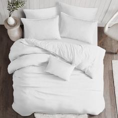 a bed with white sheets and pillows on top of wooden flooring next to a plant