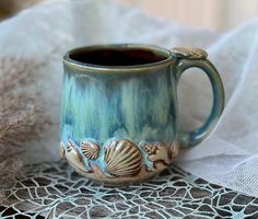 a coffee mug with sea shells on it sitting on a lace doily next to a window