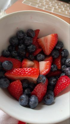 a white bowl filled with strawberries and blueberries next to a computer keyboard on a desk