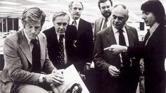 a group of men in suits and ties looking at something on a piece of paper