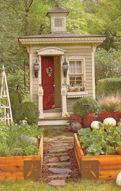 a small house with a red door surrounded by greenery and vegetables in wooden planters