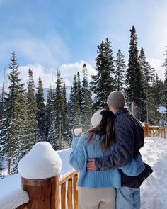 a man and woman standing on top of a snow covered slope next to pine trees