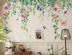 a wall with flowers and butterflies painted on the walls next to a coffee maker, lamp, and pictures