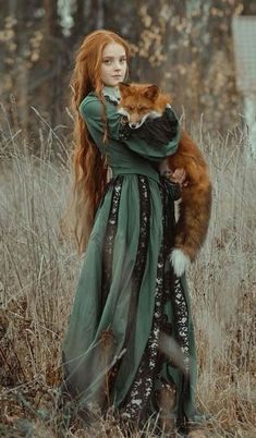 a woman with long red hair holding a cat in a grassy field next to trees