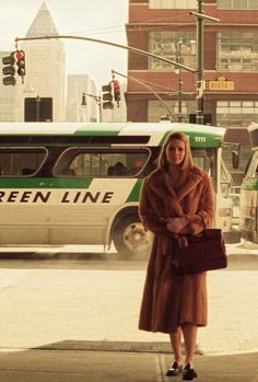 a woman in a fur coat is standing on the sidewalk near a bus and some buildings