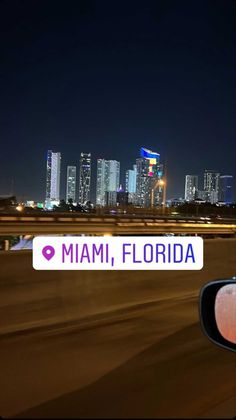 the miami skyline is lit up at night from behind a car's side view mirror