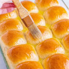 a hand holding a paint brush over a baked bread dish with rolls in the baking pan