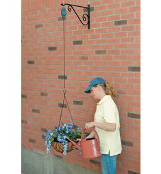 a woman holding a bucket and watering flowers on the side of a brick wall with a planter hanging from it