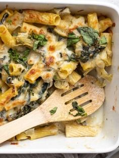 a casserole dish with spinach, cheese and pasta in it next to a wooden spoon