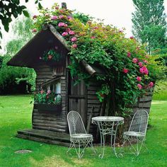 an old log cabin with flowers growing on the roof and table and chairs around it