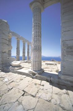 an old building with columns and stone flooring in front of the ocean on a sunny day