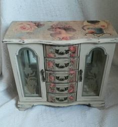 an old white cabinet with pink roses on the top and glass doors, is sitting on a bed