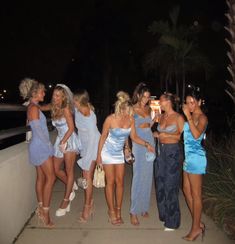 a group of young women standing next to each other on a sidewalk at night with palm trees in the background