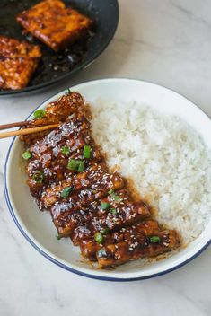 some meat and rice on a plate with chopsticks next to it in a bowl