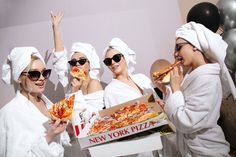 three women in white robes are eating pizza and posing for the camera with their hands up