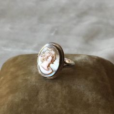 This is such a beautiful ring! It has an oval shaped carved cameo of a woman in profile, with a carved necklace on her neck and shoulder length hair. The cameo looks to be carved of mother of pearl or shell. The cameo is pink with a soft toned blue grey background Maker: Unknown Age: Unknown, estimated to between 1900-1940 Color: Silver, peach pink, blue grey Material: The metal is unmarked but acid tests to sterling. Cameo is shell or mother of pearl, carved. Condition: There's a small area of Elegant Oval Cameo Jewelry, Oval Mother Of Pearl Ring With Polished Finish, Oval Mother Of Pearl Jewelry With Polished Finish, Formal Oval Mother Of Pearl Rings, Classic Oval Cameo Jewelry, Round Intaglio Jewelry, White Oval Cameo Jewelry, Heirloom Oval Cameo Jewelry, Elegant Cameo Rings