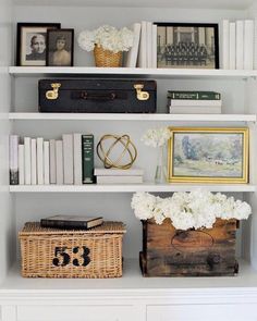 a white bookcase with books and pictures on it, along with flowers in a basket