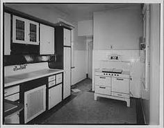 an old black and white photo of a kitchen with stove, sink, cabinets and cupboards