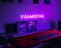 two computer monitors sitting on top of a wooden desk in front of a neon sign