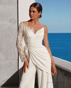 a woman in a white dress standing next to a wall with the ocean behind her