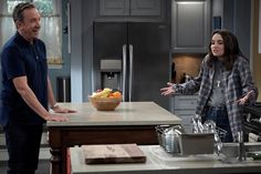 two people standing in a kitchen talking to each other and one person holding a bowl of fruit