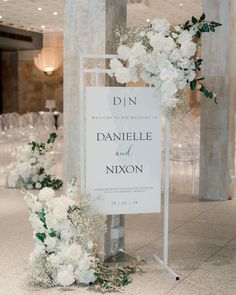 a sign with flowers and greenery on it in the middle of a room filled with chairs