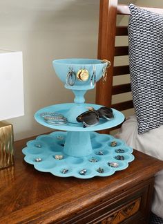 two tiered tray with sunglasses and jewelry on it in front of a bed, next to a night stand