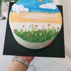 a hand holding up a cake decorated with grass and flowers in the sky on top of it