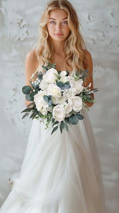 a woman in a white wedding dress holding a bouquet of flowers and greenery on her shoulder