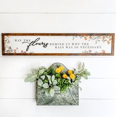 a sign hanging on the side of a wall with flowers and plants in front of it