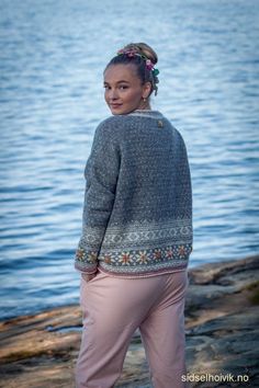 a woman standing on rocks near the water wearing pink pants and a sweater with an intricate design