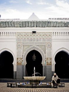 two people sitting on benches in front of a building with a fountain and tiled roof