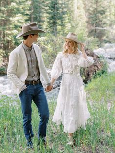 a man and woman in cowboy hats walking through tall grass next to a river holding hands