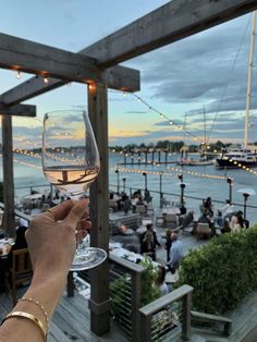 a person holding up a wine glass in front of an outdoor dining area with boats on the water