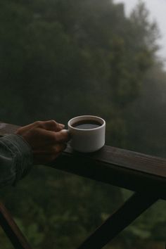 a person holding a cup of coffee on top of a wooden table next to a forest