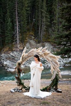 a woman standing in front of a river holding a large circular wreath with feathers on it